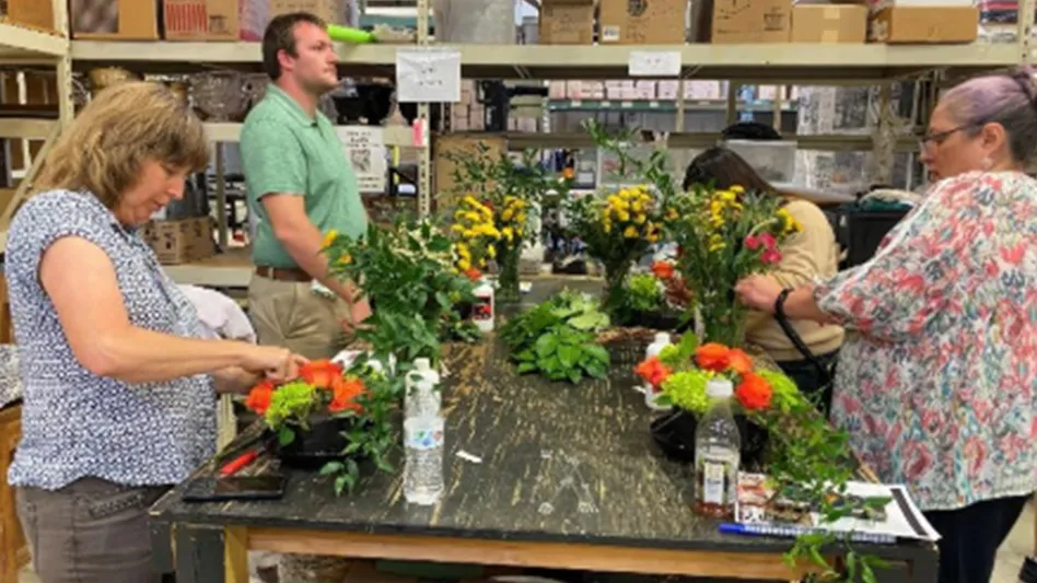 Four people work on flower arrangements on a black table in a room with brown cardboard boxes on shelves behind them.